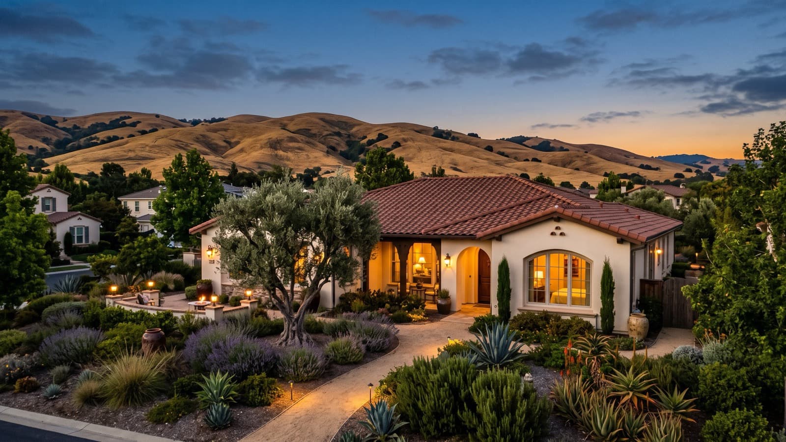 A single-story Spanish-style home with a red tile roof and lit windows at dusk, framed by an olive tree and drought-tolerant garden, with golden hills behind it.